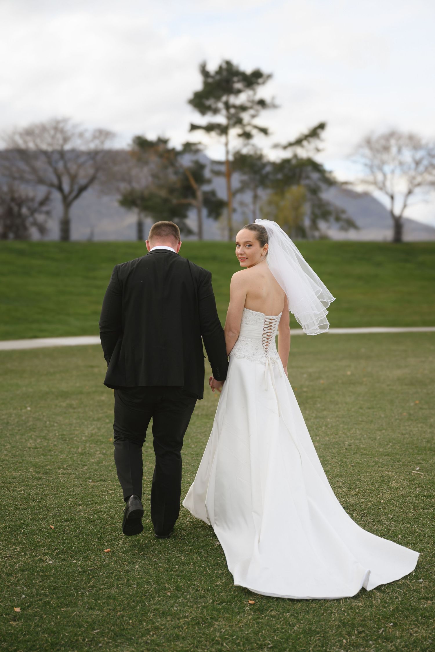 Bride in white strapless gown with veil looks back while walking with groom on green lawn.