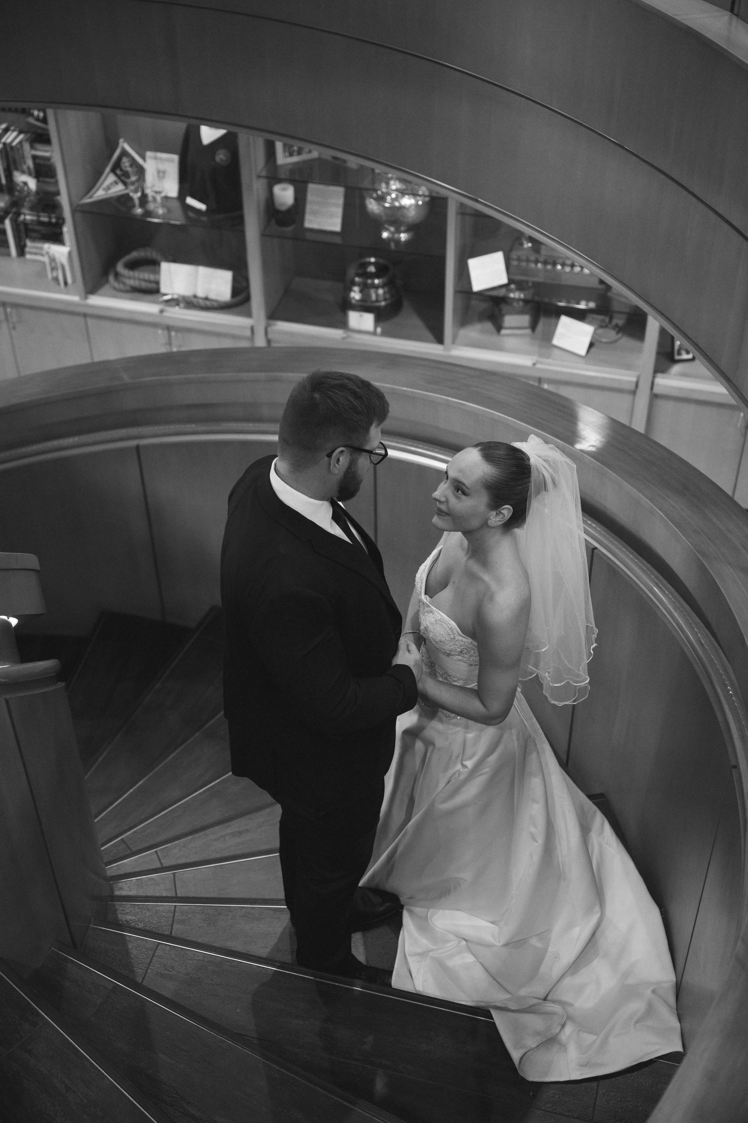 Black and white photo of bride and groom holding hands on spiral staircase, bride in white gown and veil.