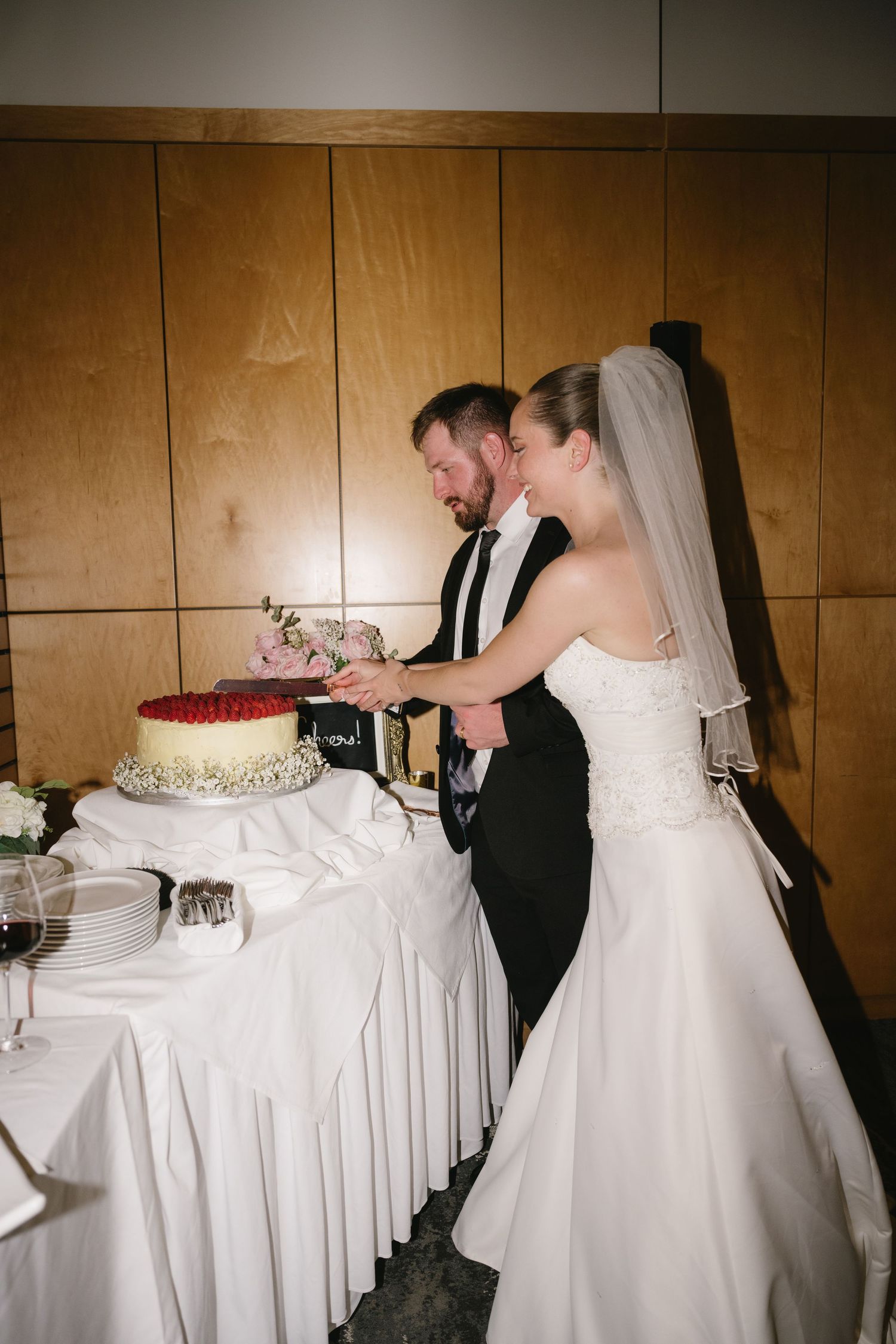 Bride and groom cutting wedding cake together at reception, bride in strapless lace gown with veil.