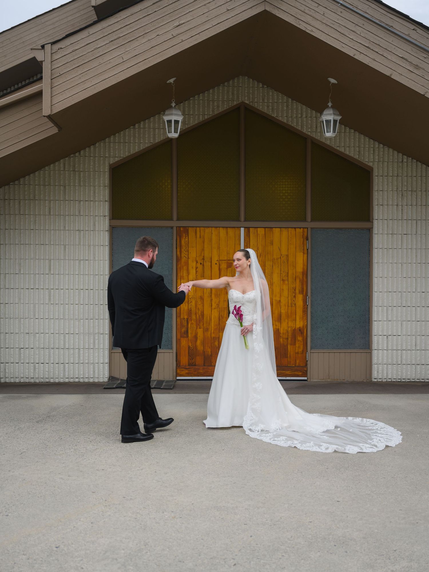 Bride in long white gown with cathedral veil holding groom's hand outside church entrance.