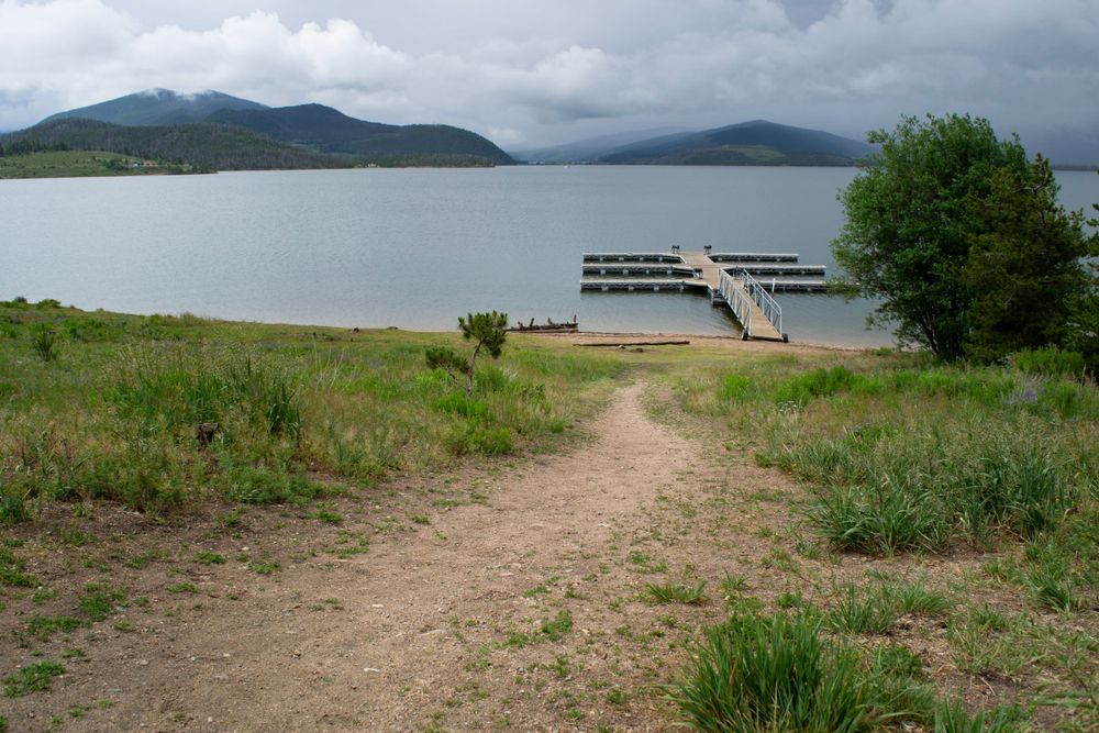 A dirt path leads down to a wooden dock on a serene lake surrounded by mountains under cloudy skies.