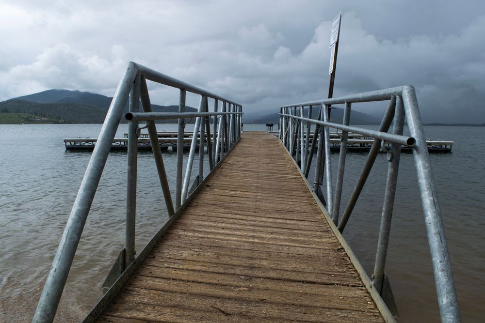Long wooden pier with metal railings stretches out over calm lake waters with mountains in the distance.