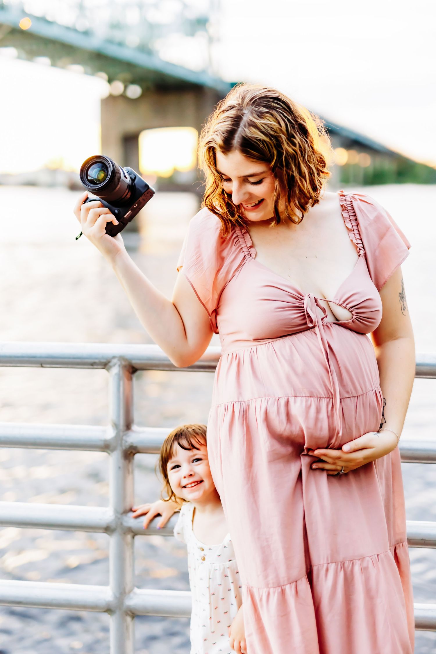 pregnant mama holding her camera with her toddler daughter smiling behind her, on the river under the cape fear memorial