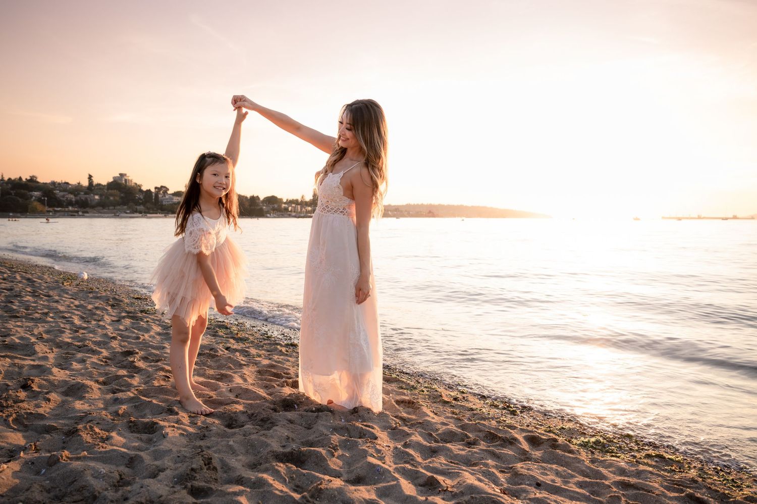 Two people in white dresses dance together on a sandy beach at sunset with ocean waves in the background.