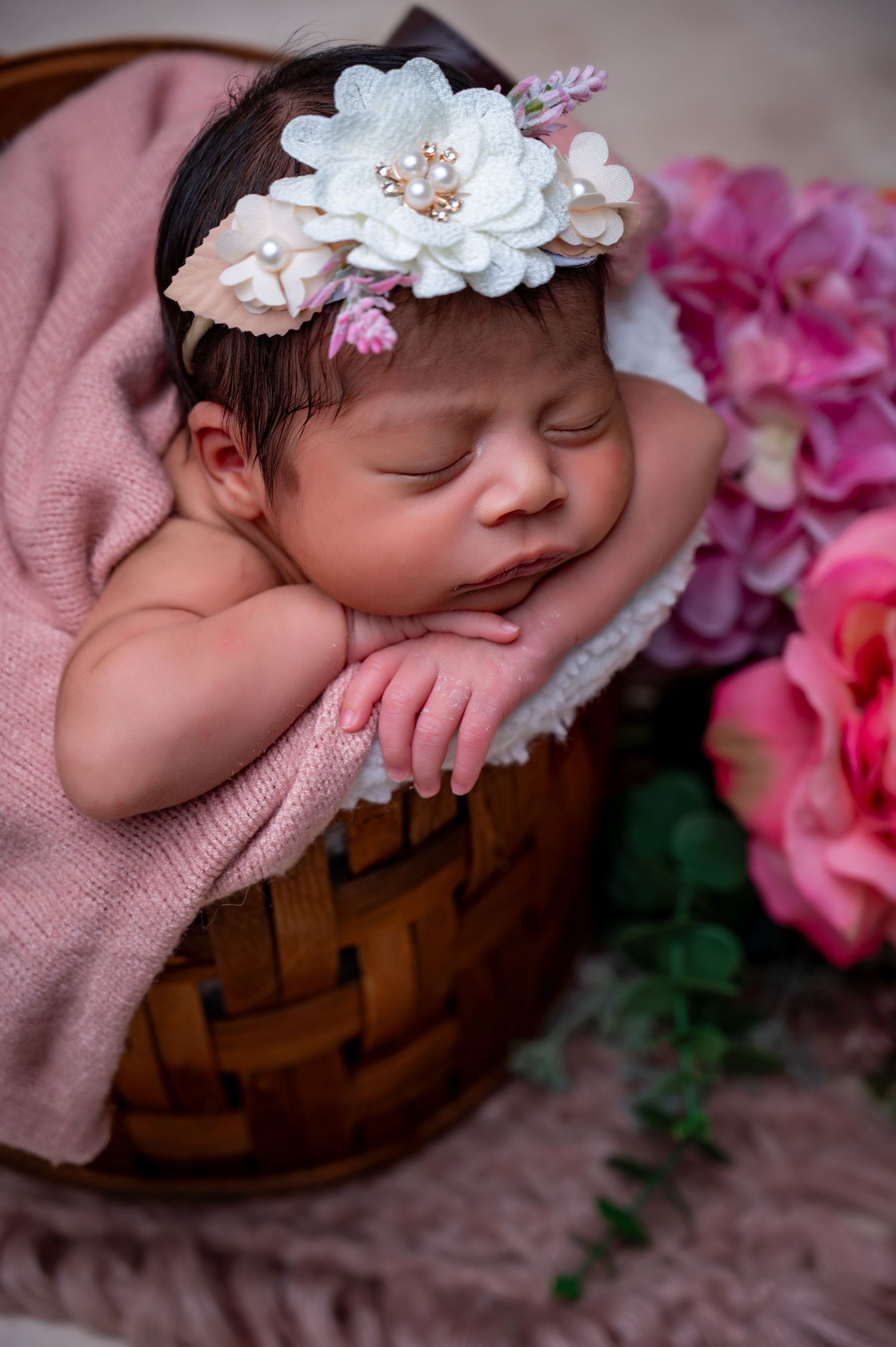 Newborn baby resting in wooden basket with pink flowers and white floral headband.