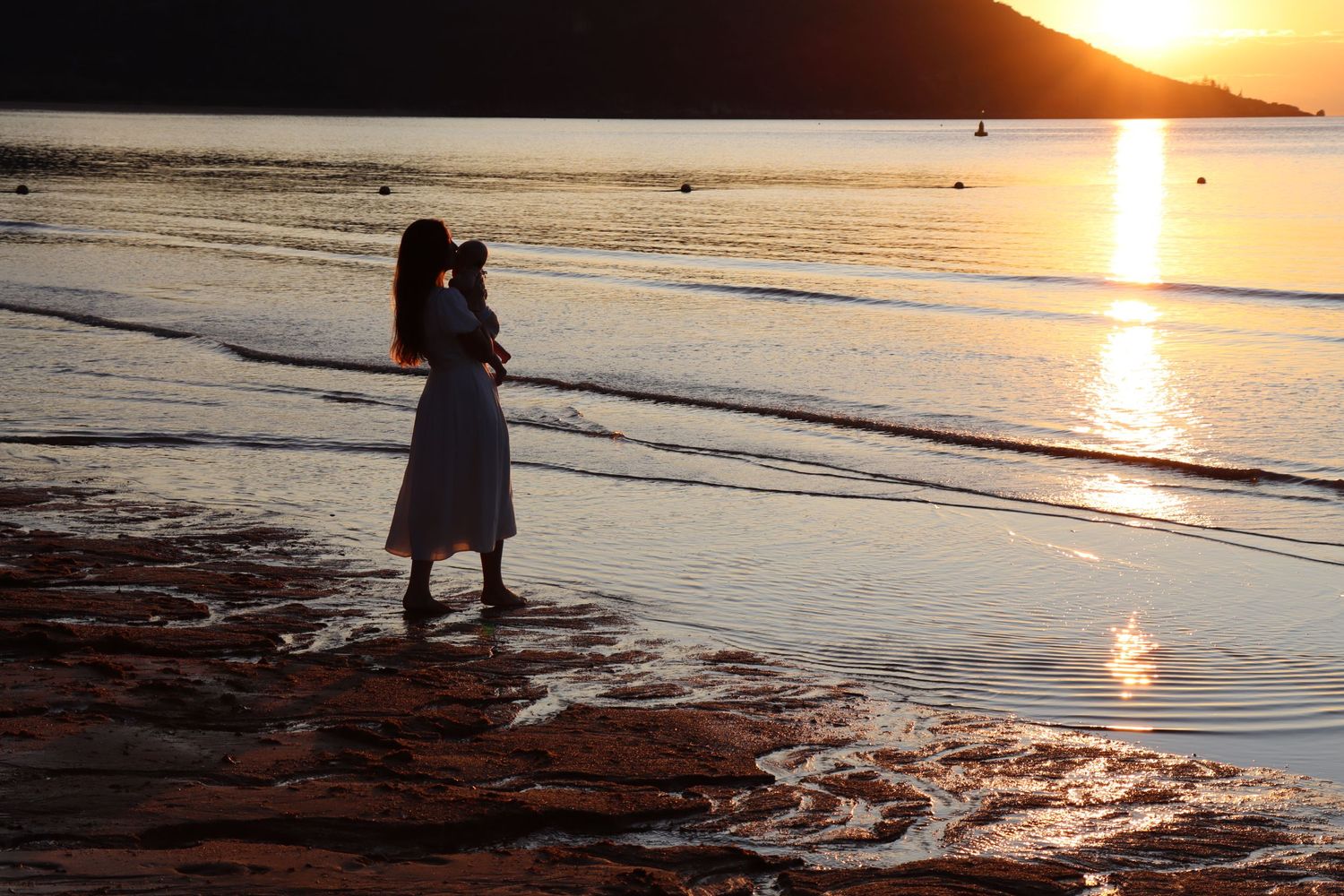 Woman in white dress standing on a rocky beach at golden sunset with calm ocean waters reflecting warm light.