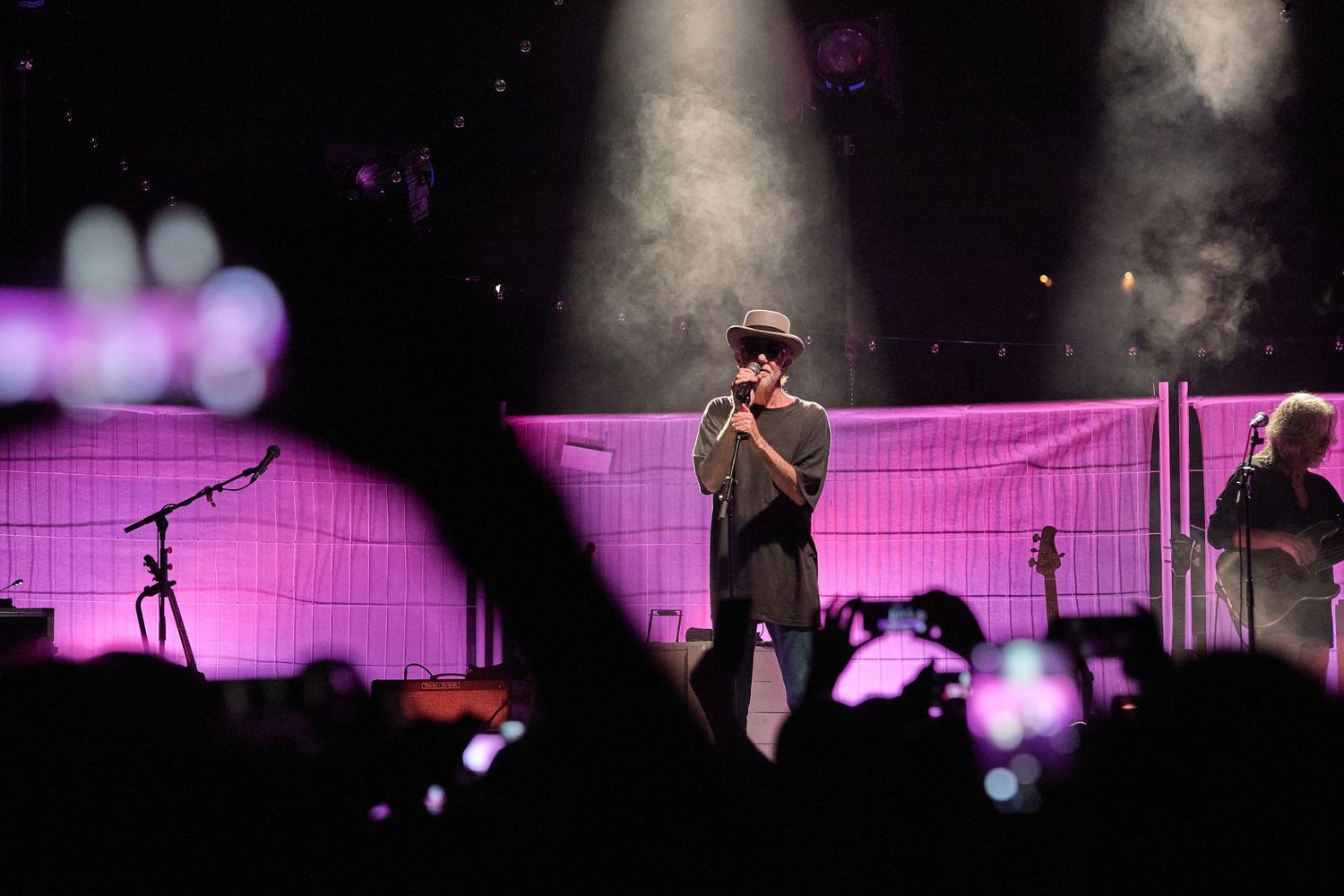 Concert performer on stage illuminated by spotlight with purple backdrop and crowd phones visible.