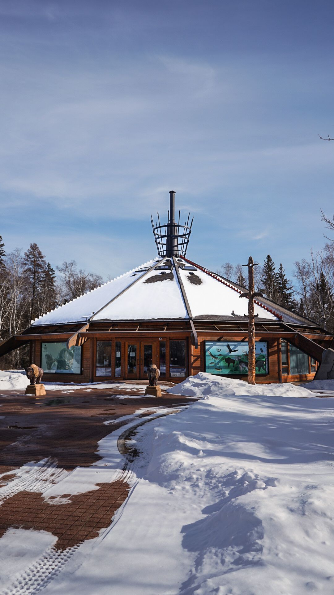 Snow-covered log cabin lodge with wooden deck and tall windows against a bright blue winter sky.