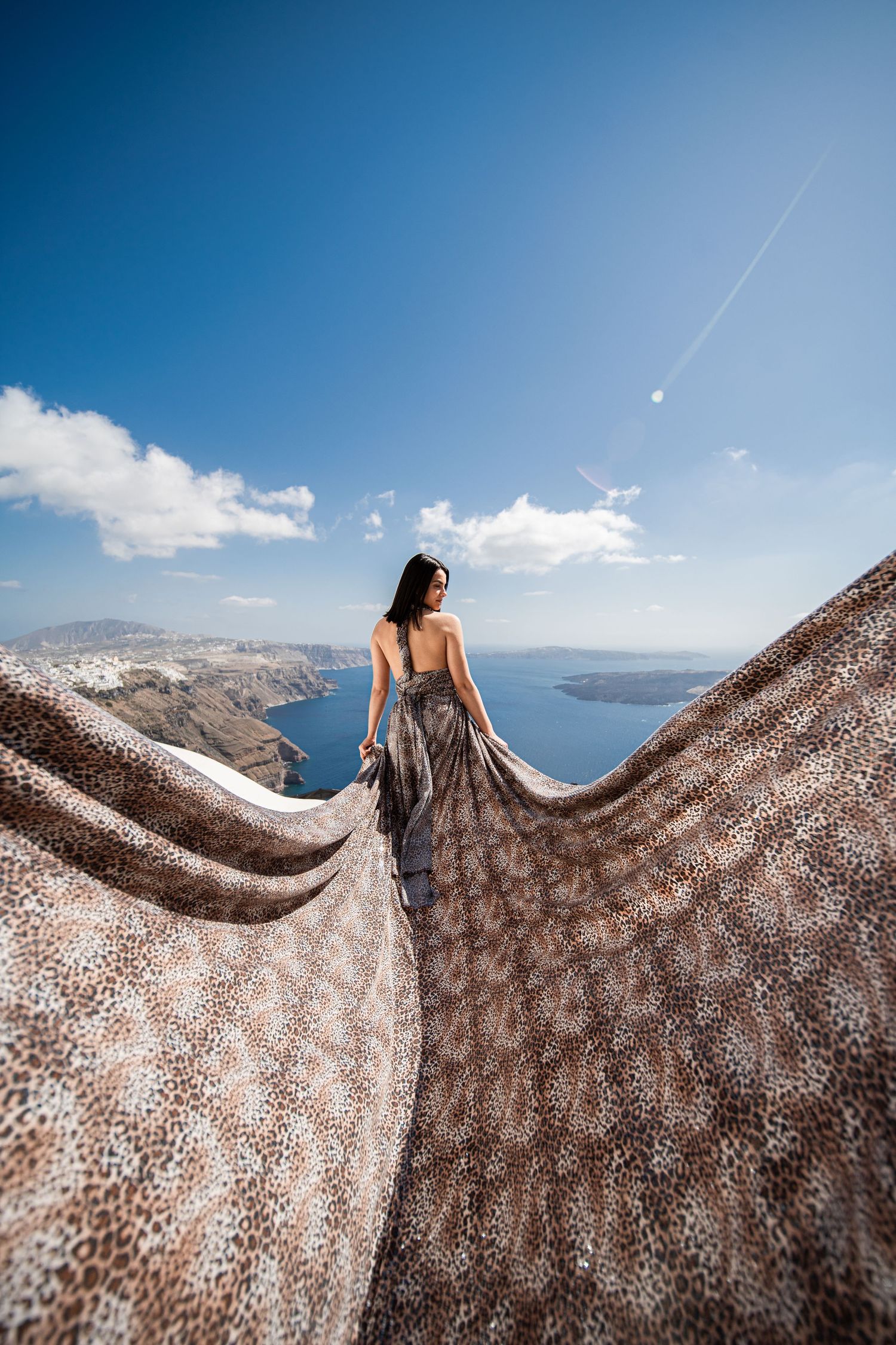 Patterned fabric dress spreads dramatically against Santorini's iconic coastal landscape.