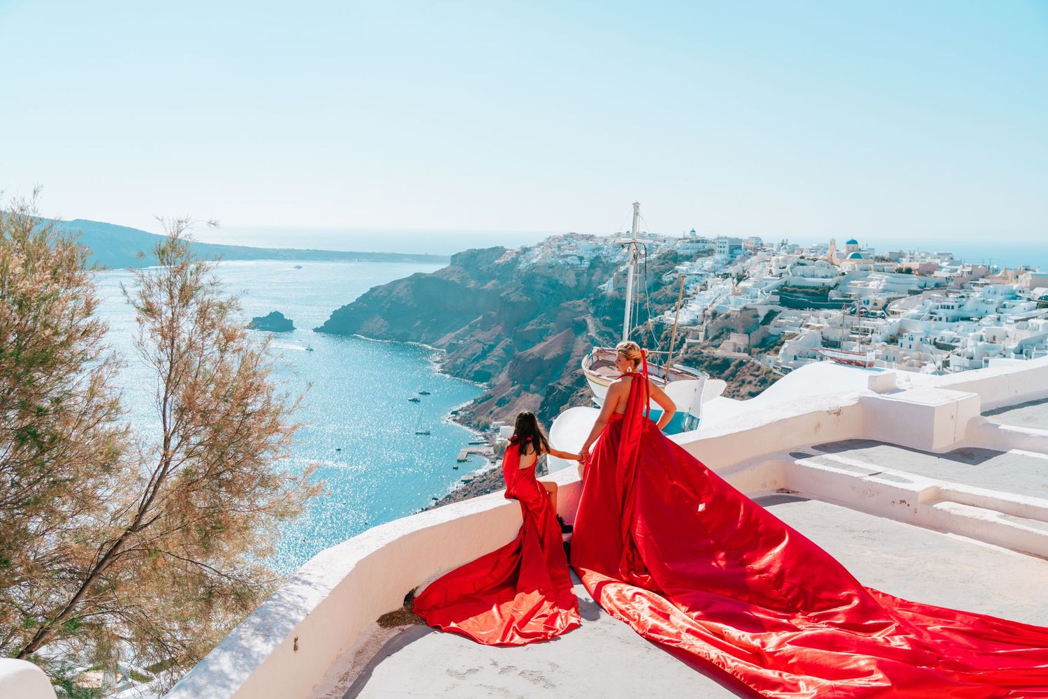 Red flowing dresses create dramatic scene on white terrace overlooking Santorini's coast.