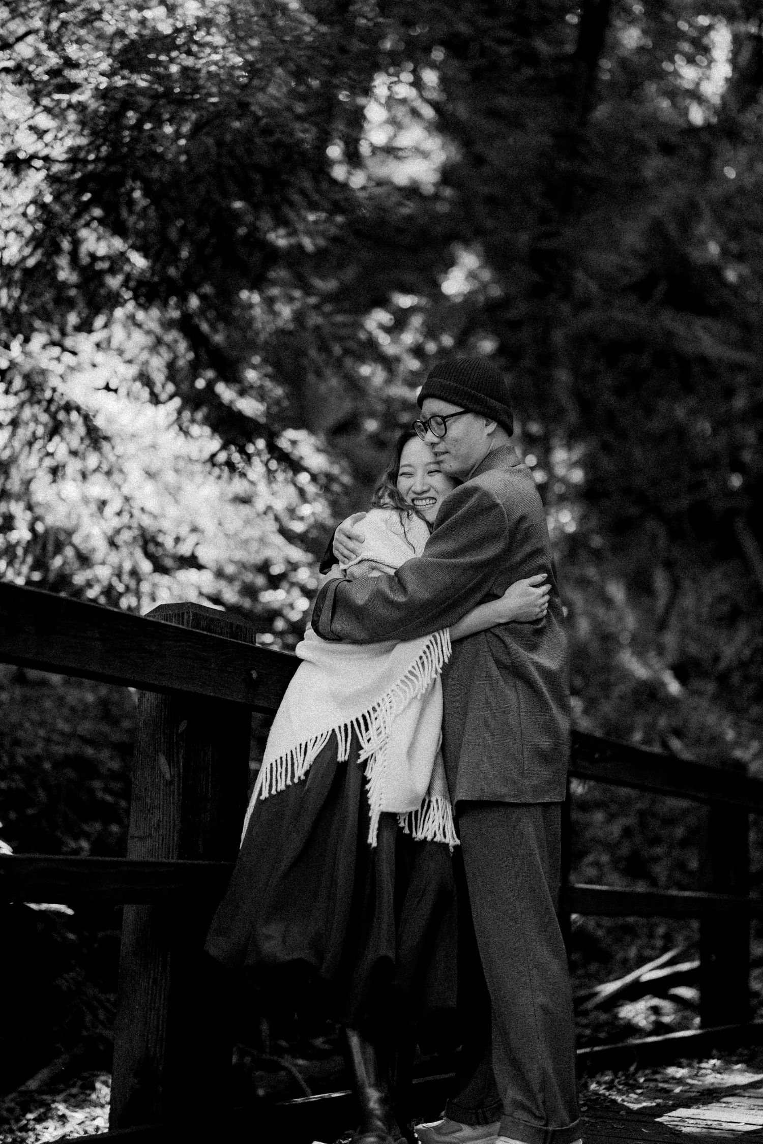 Black and white photo of a couple embracing on a wooden bridge in a forest setting.