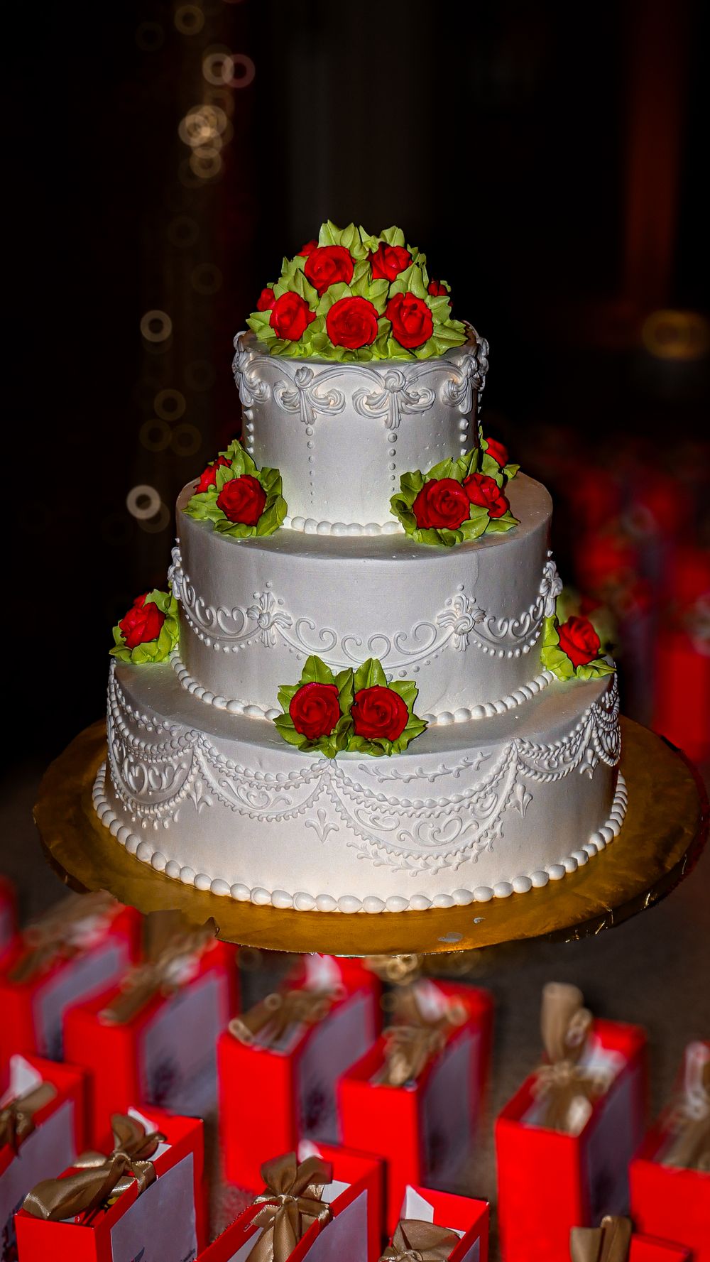 Elegant three-tier white wedding cake decorated with red roses and intricate piping details on a gold cake stand.