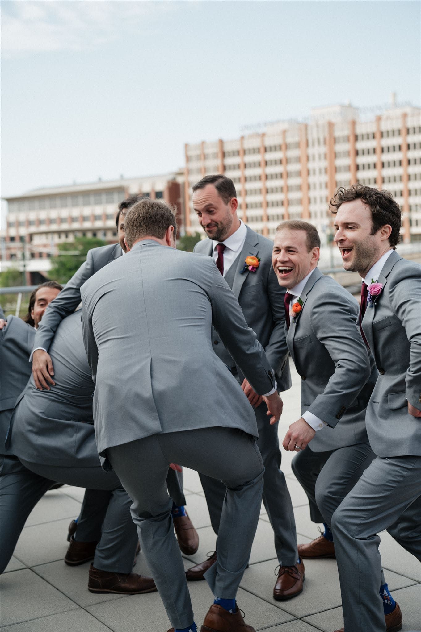 Groomsmen in matching gray suits laughing and dancing together at an outdoor wedding celebration.
