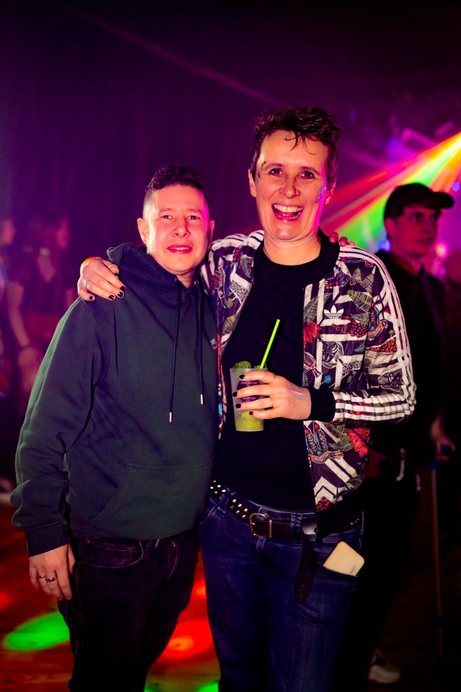 Two friends pose together at a nightclub with colorful lights and neon effects in the background.