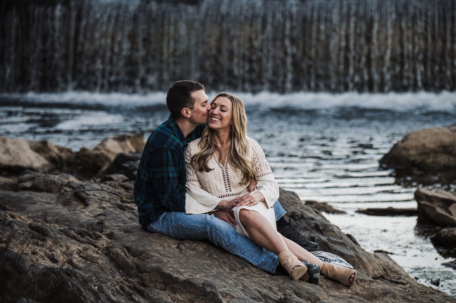 Romantic couple embracing on rocky shore near waterfall.