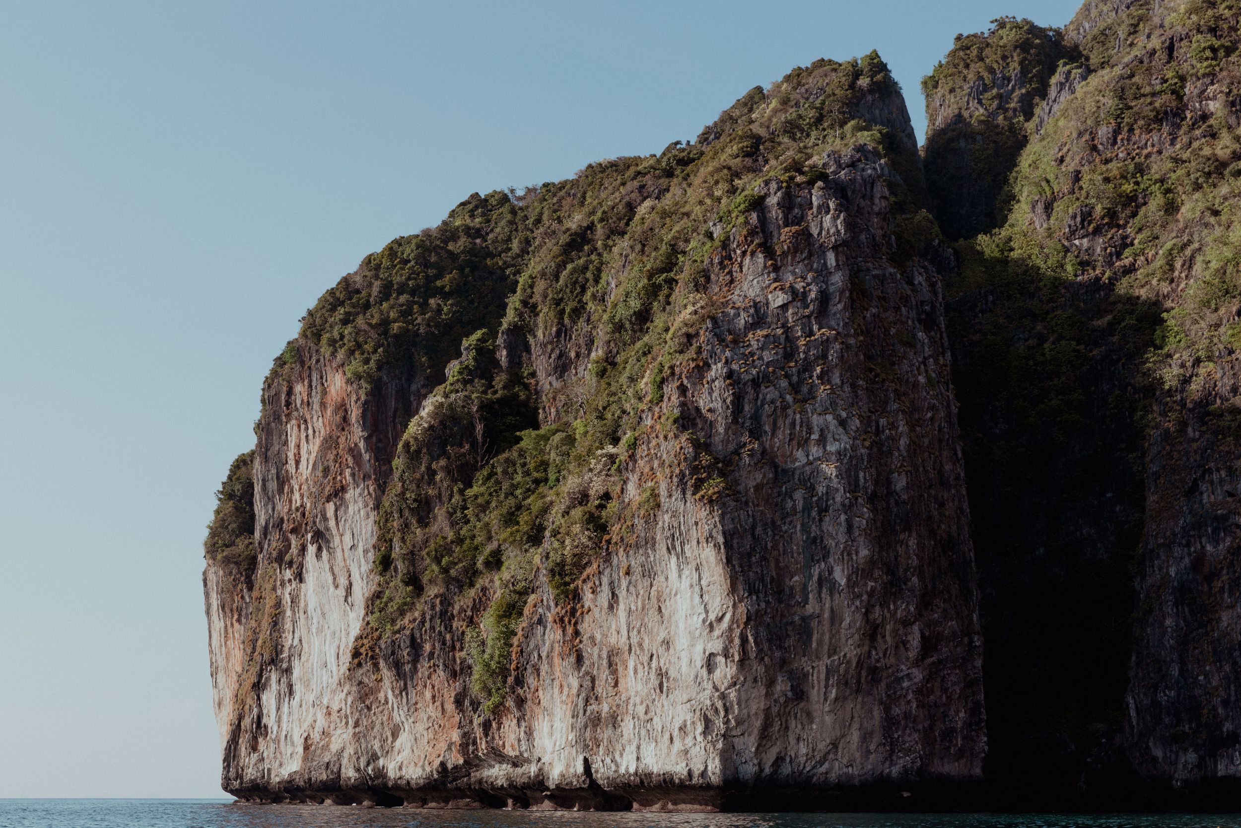 Towering limestone cliff rising from the ocean with vegetation growing on its face.