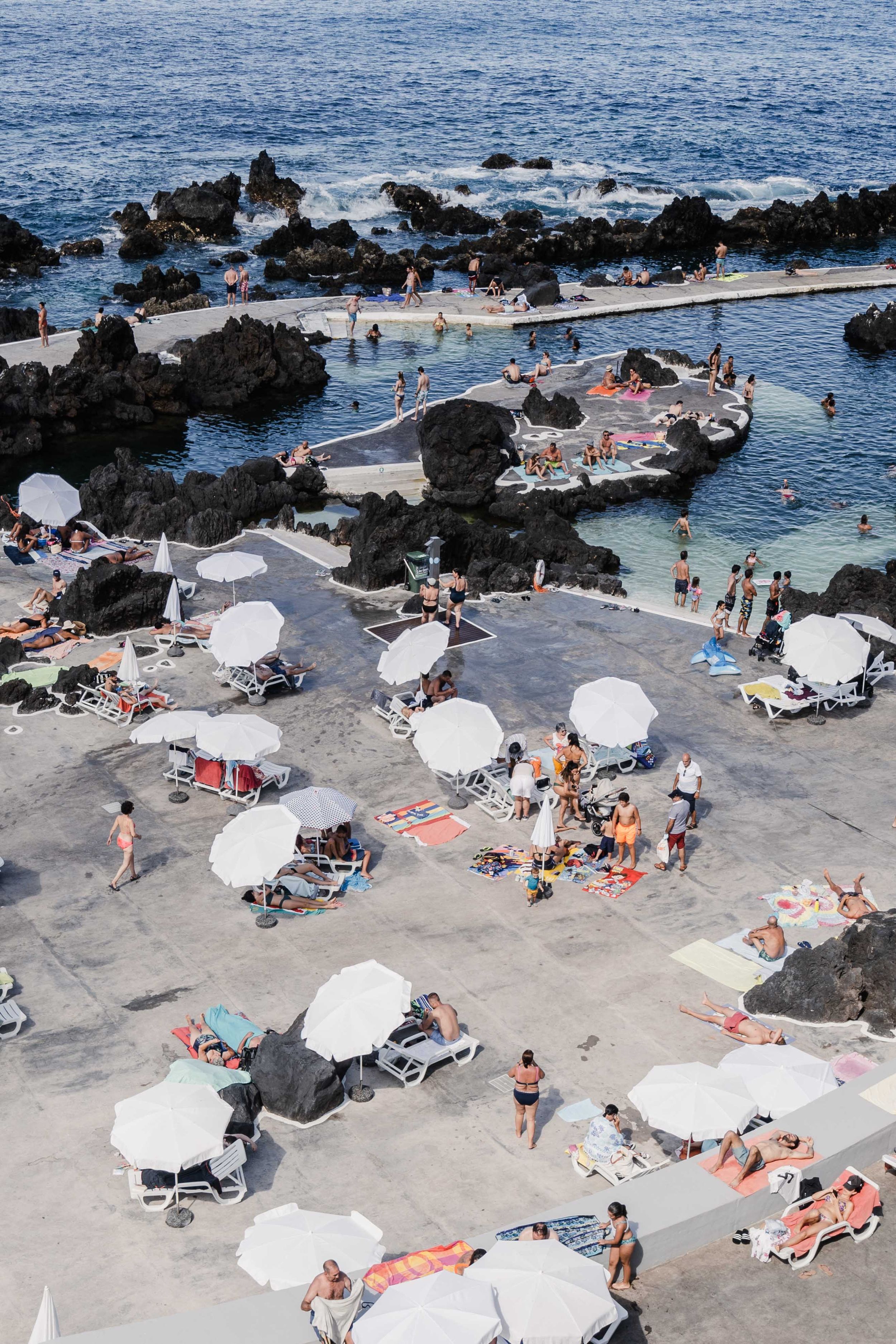 Natural rock pool swimming area with white umbrellas and sunbathers along the rocky coast.