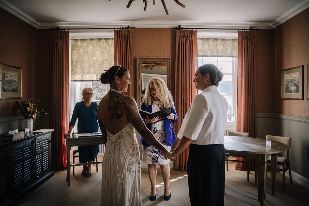 Bride in backless wedding dress holds hands with guest during intimate indoor wedding ceremony with officiant.