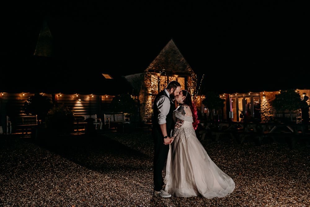 Couple in wedding attire dancing together outside a rustic venue at dusk with twinkling lights overhead.