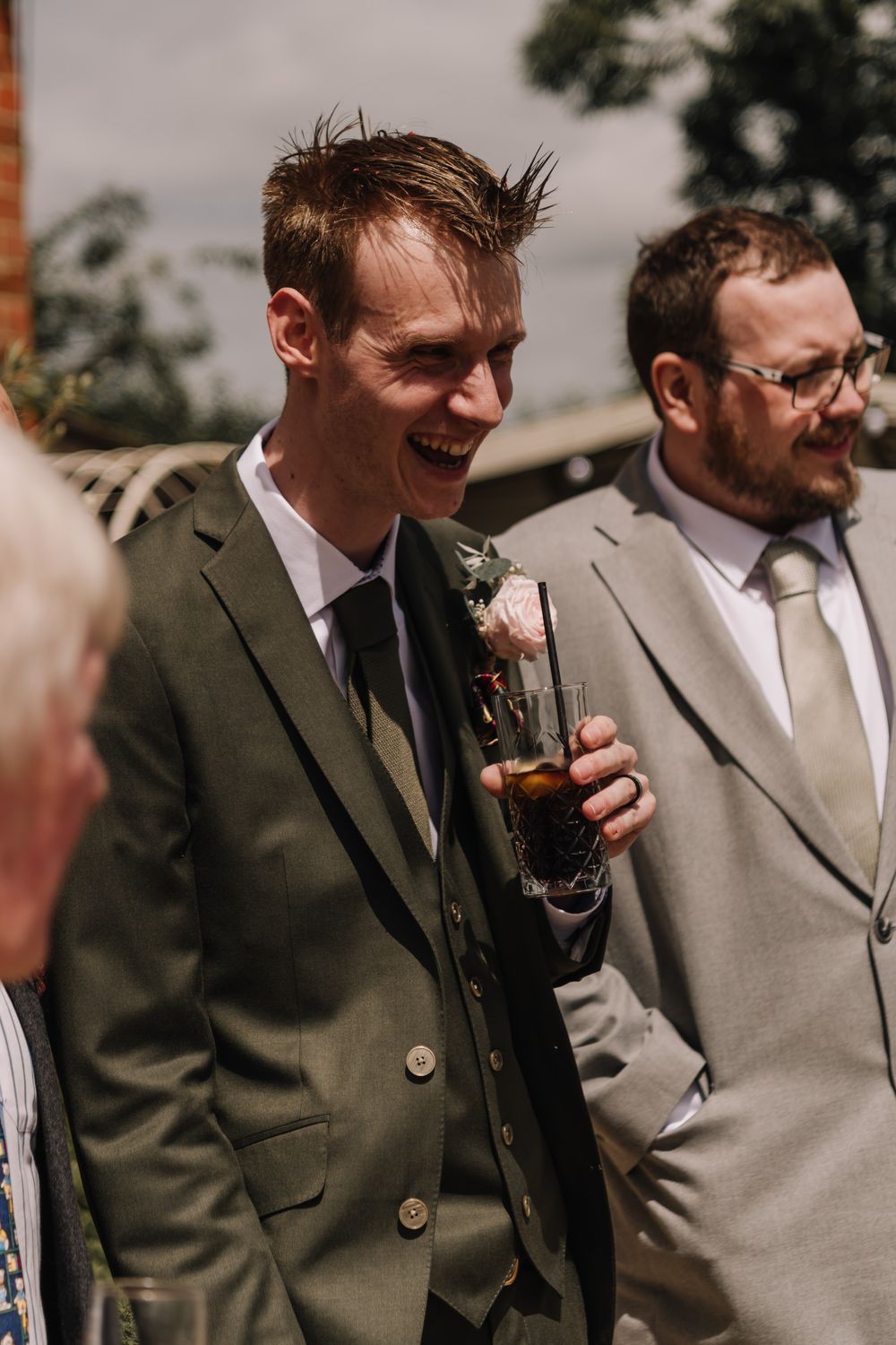 Groom laughing in green suit with boutonniere, holding drink at outdoor wedding reception.
