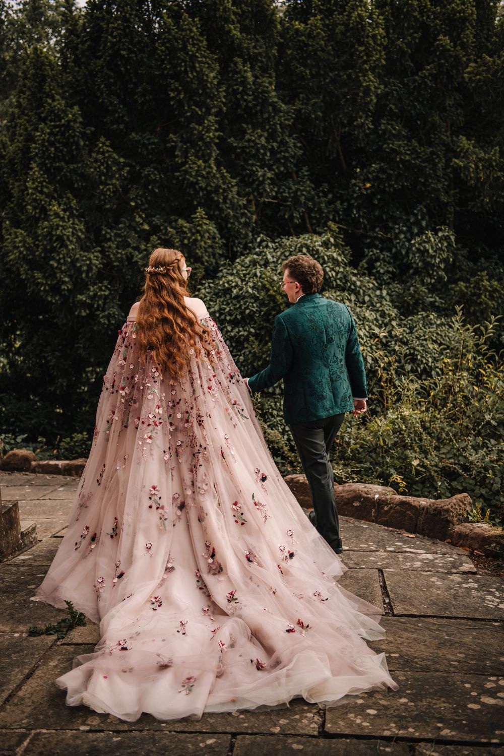 Bride in pink floral cape gown holds hands with groom in teal suit, walking through lush garden.