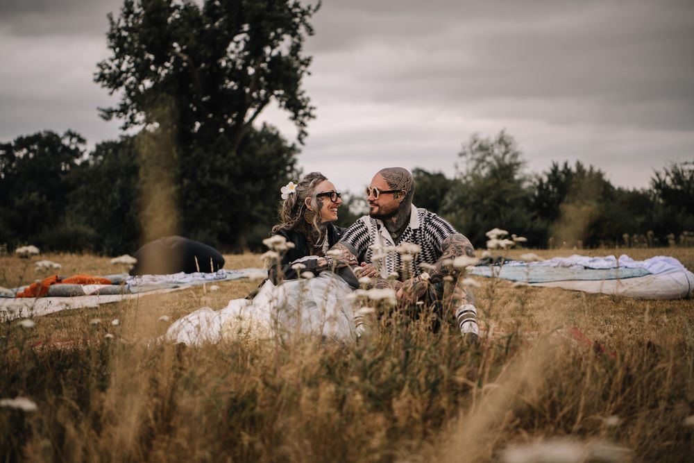 Couple sitting together in a golden meadow on a cloudy day, surrounded by wildflowers and a picnic blanket.