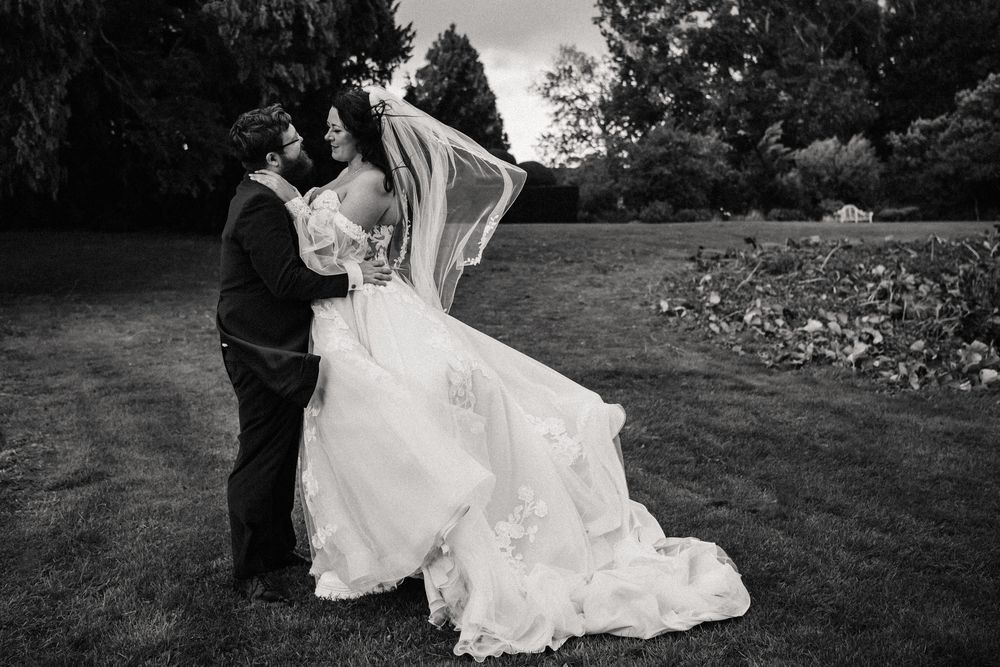 A romantic wedding couple embraces in a garden setting while the bride's white dress flows in the breeze.
