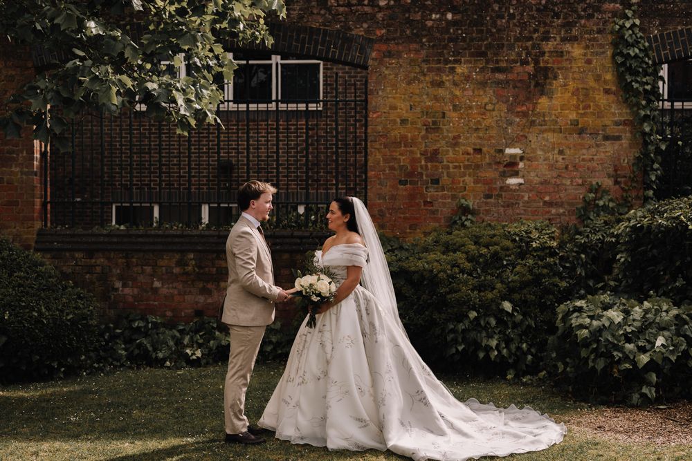 A bride in a white gown and groom in a tan suit hold hands in a lush garden beside a brick wall.