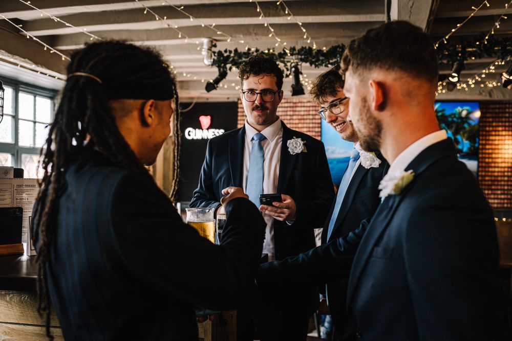 Groom and wedding guests socializing at a rustic venue bar with fairy lights and Guinness signage.