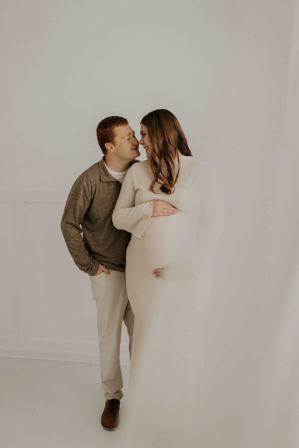 A couple shares an intimate moment during a minimalist maternity photoshoot against a white backdrop.