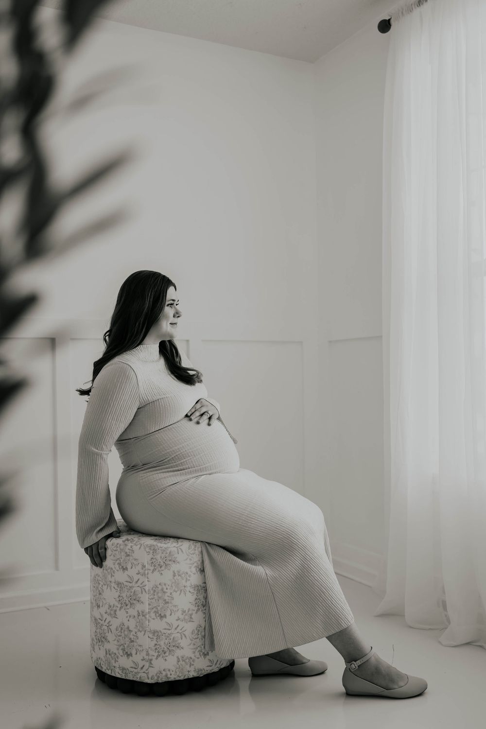 A series of black and white maternity photos showing a pregnant woman in a fitted dress posing against a white wall.