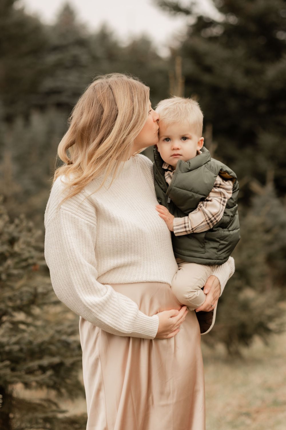 Sweet outdoor maternity photo featuring a woman in a cream sweater holding a toddler in a green vest.