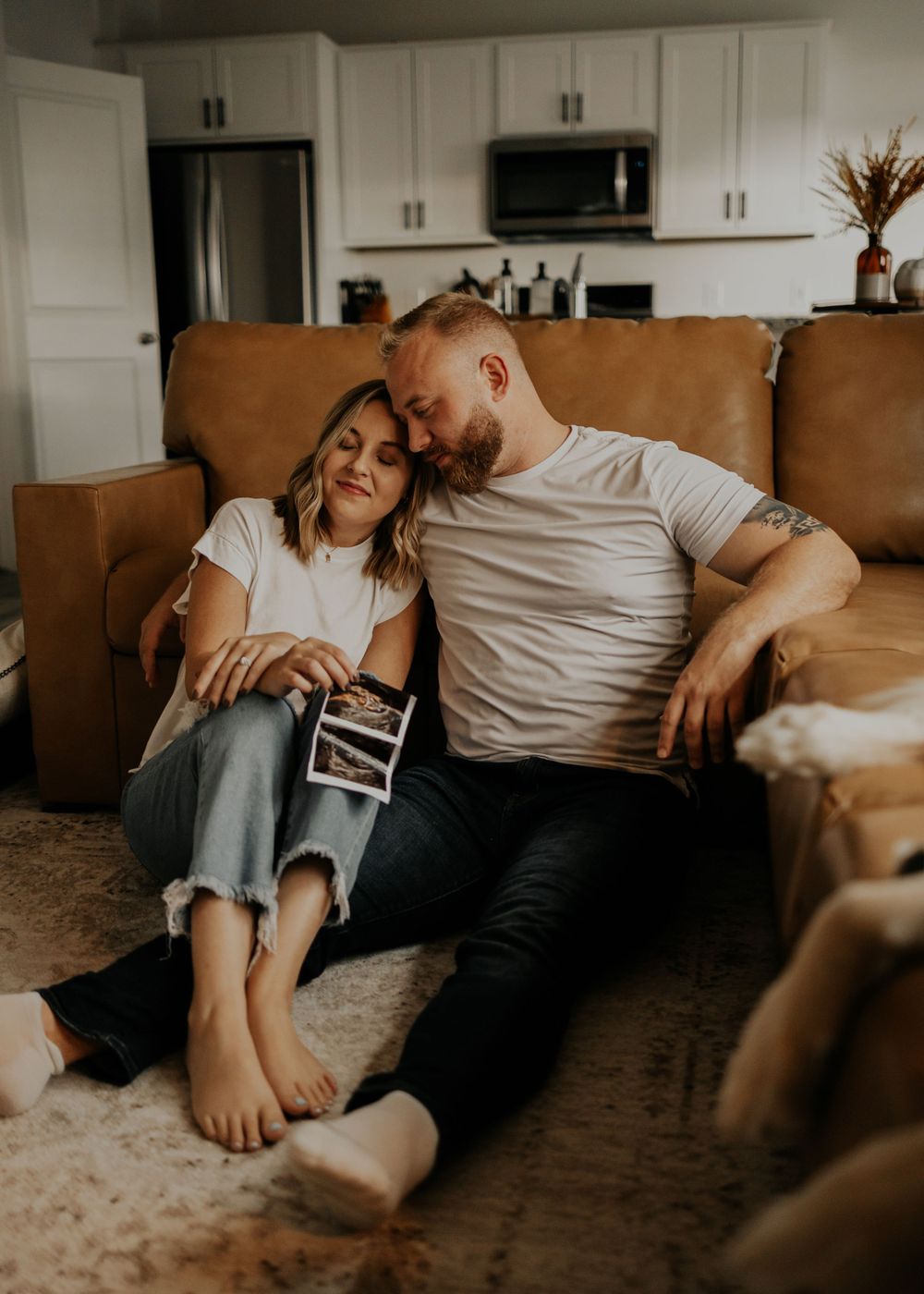 A couple sits together on a brown leather couch holding an ultrasound photo in their living room.