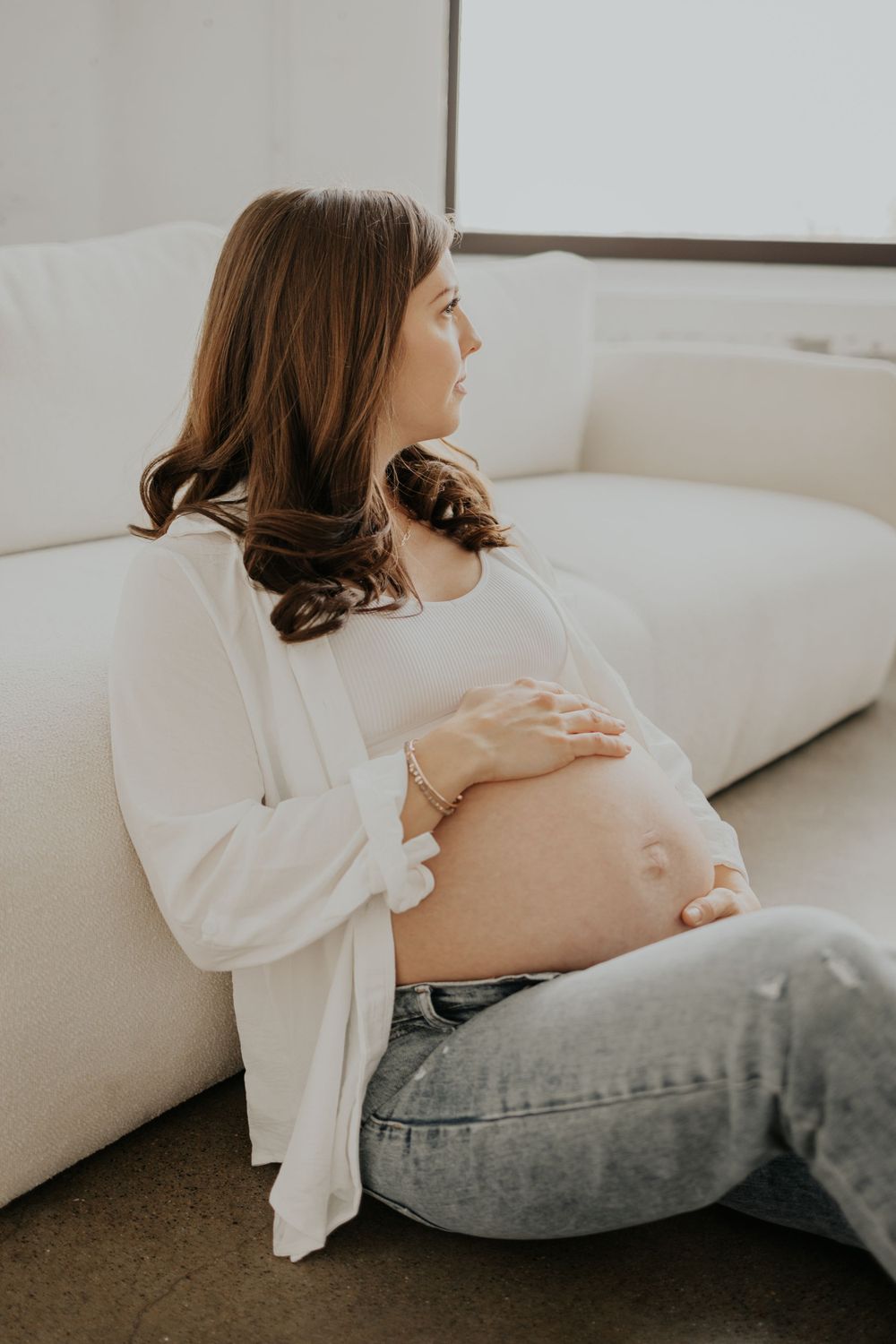 Side view of an expectant mother in white sweater and jeans sitting on white couch while cradling her baby bump.