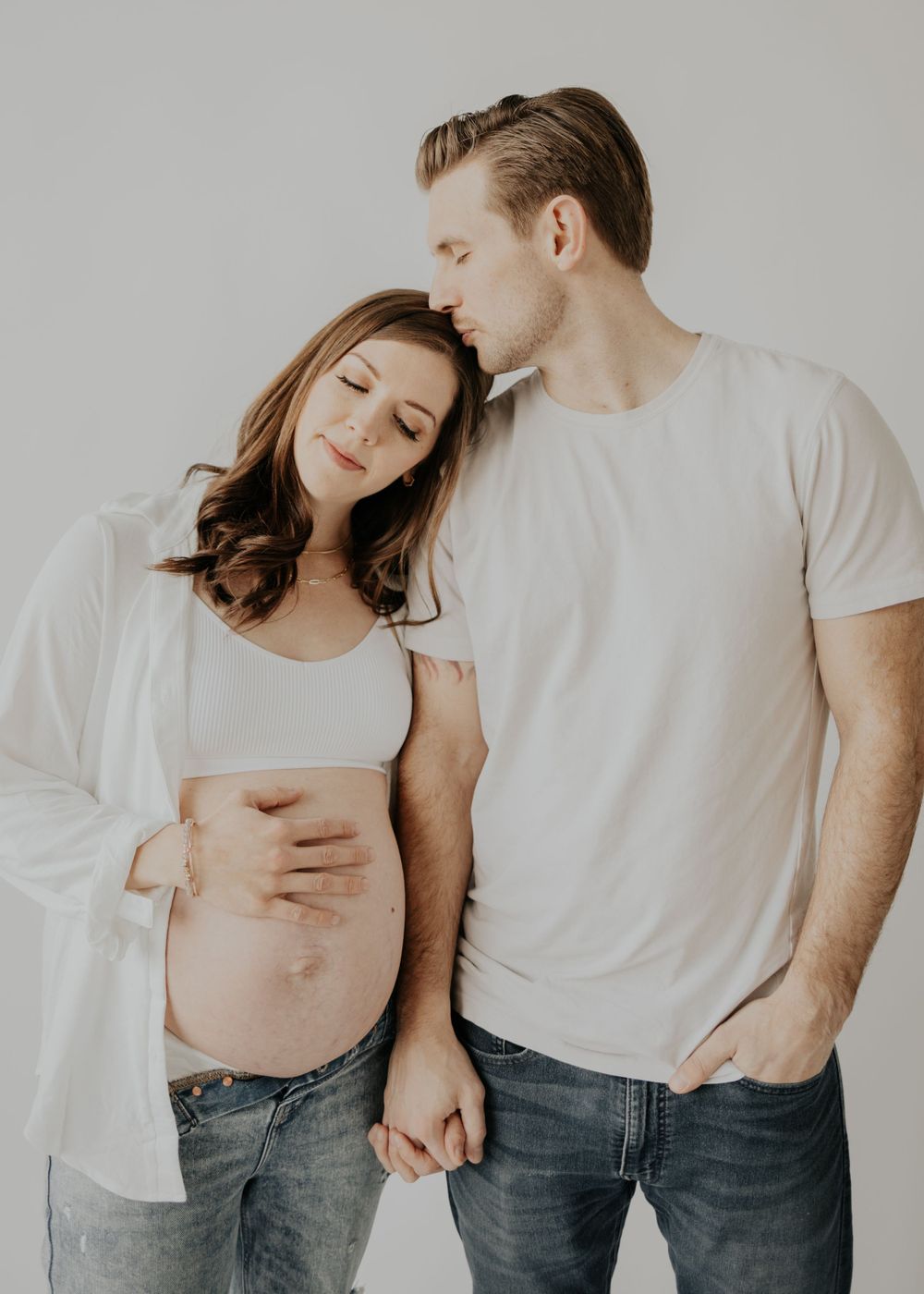 A tender maternity photo features an intimate moment between a couple in white shirts and jeans against a light backdrop.