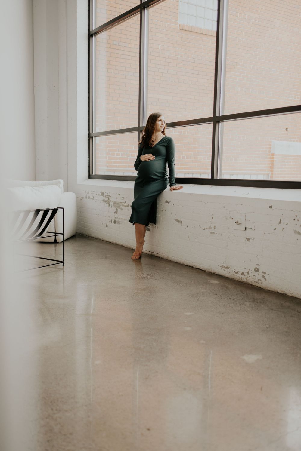 Expectant mother in emerald green dress stands contemplatively by large industrial windows in minimalist concrete space.