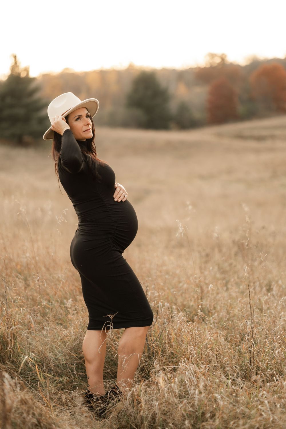 Expectant mother in black dress and white hat poses in golden field during sunset.