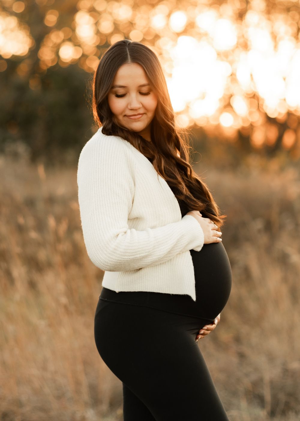 An expectant person in a white sweater poses during golden hour sunset in a field of dry grass.