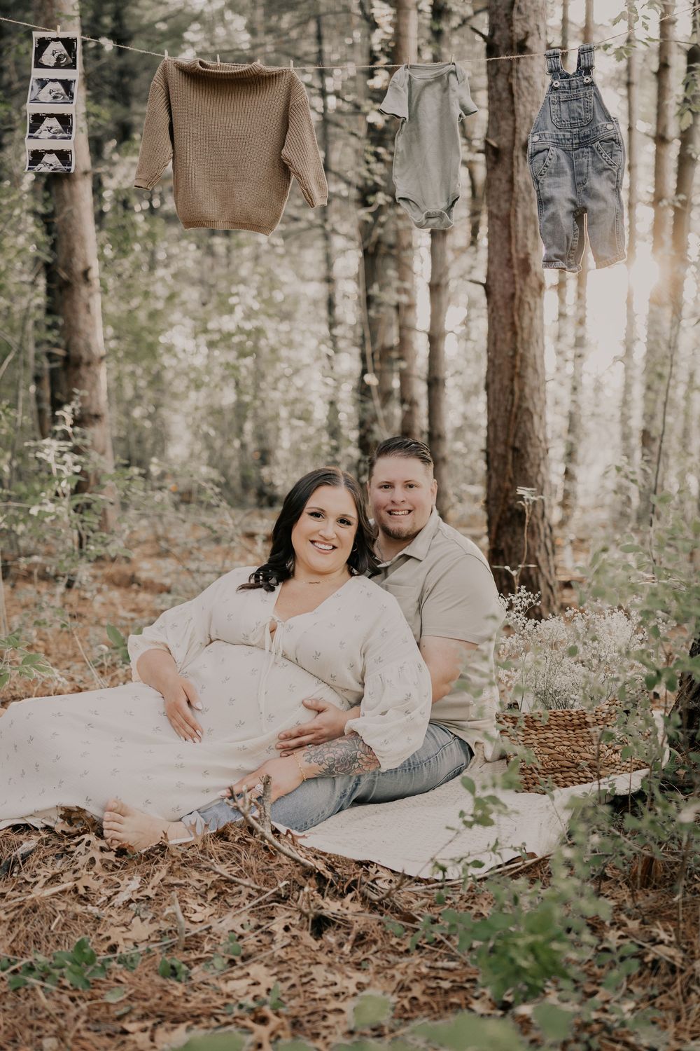 A maternity photo session in a forest setting with clothes hanging on trees and a couple sitting on a blanket in soft light.