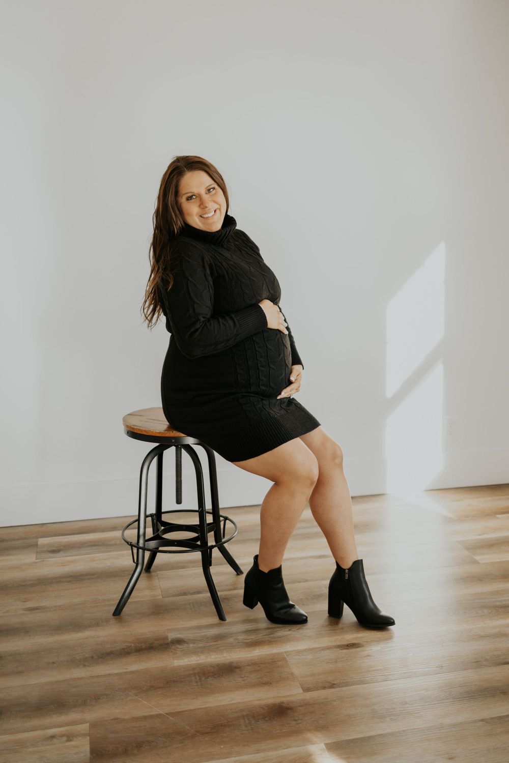 An expectant woman in a black dress and ankle boots sits on a wooden stool against a white wall.