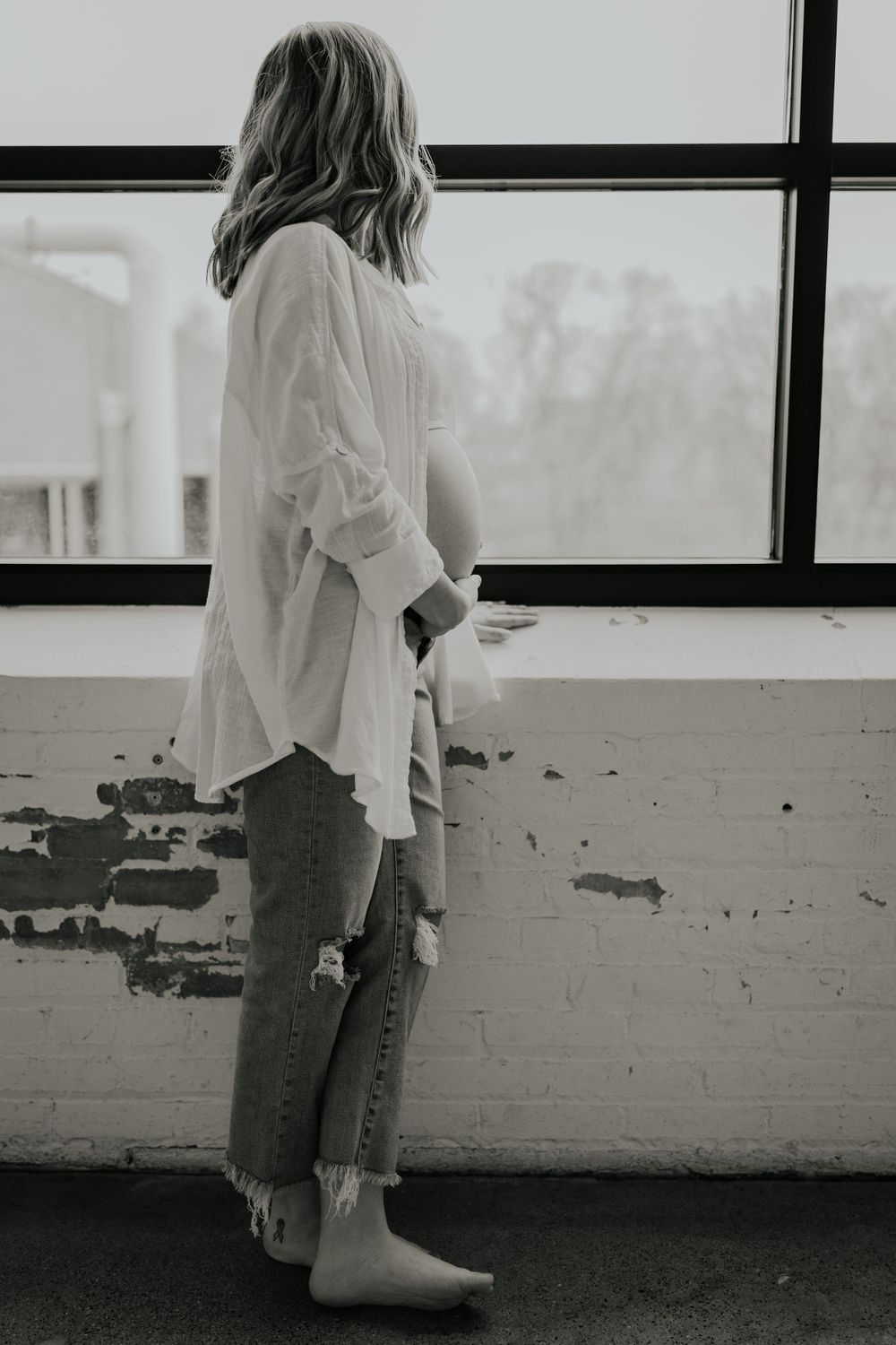 A person in a white button-down shirt and cropped jeans stands by a window in black and white photography.