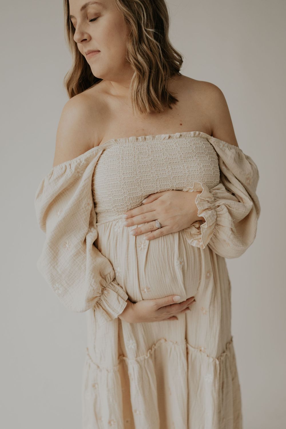 A minimalist maternity portrait features an off-shoulder cream dress with smocked bodice and billowy sleeves against a neutral backdrop.