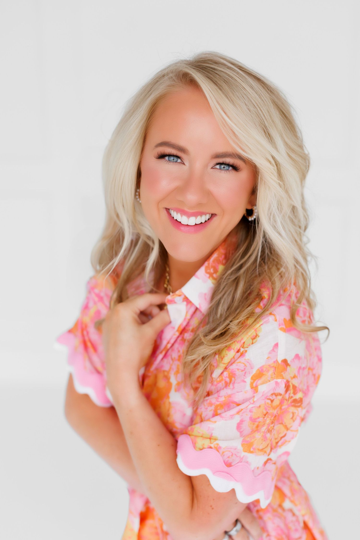 Professional lifestyle portrait of a blonde woman in a white blazer sitting on a gray couch smiling against light background.