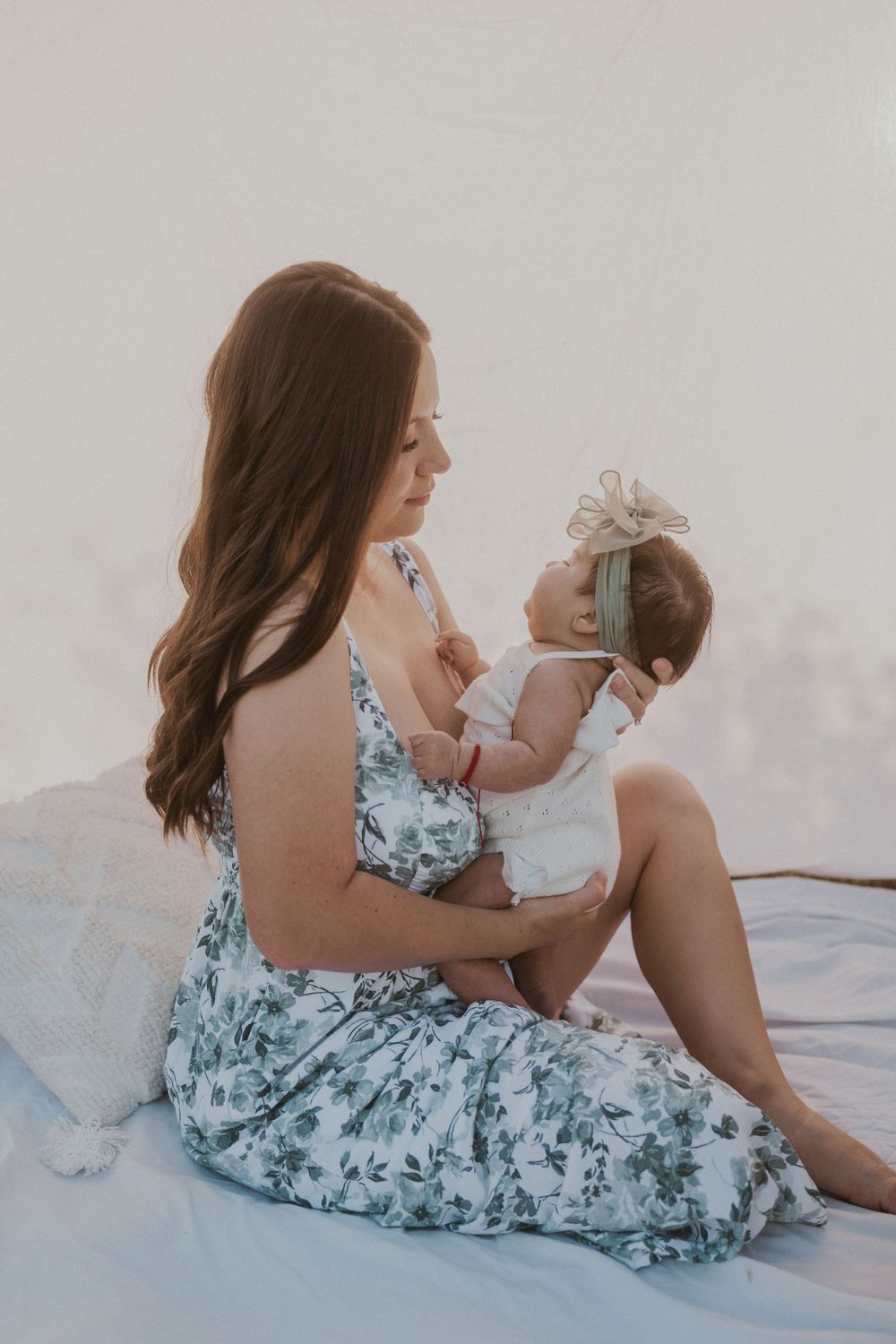 Woman in floral dress holds baby while sitting on white blanket with soft lighting.