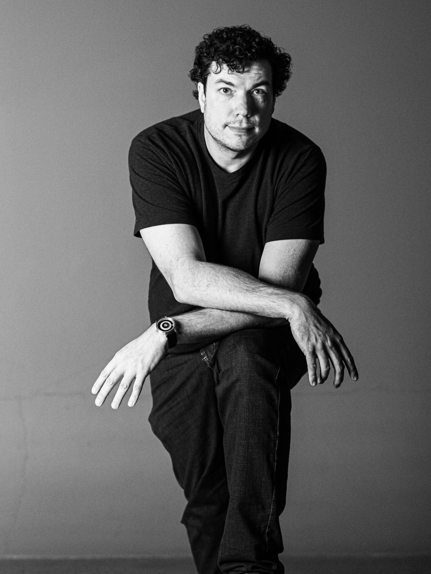 Black and white studio portrait of a person in dark clothing sitting against grey background.