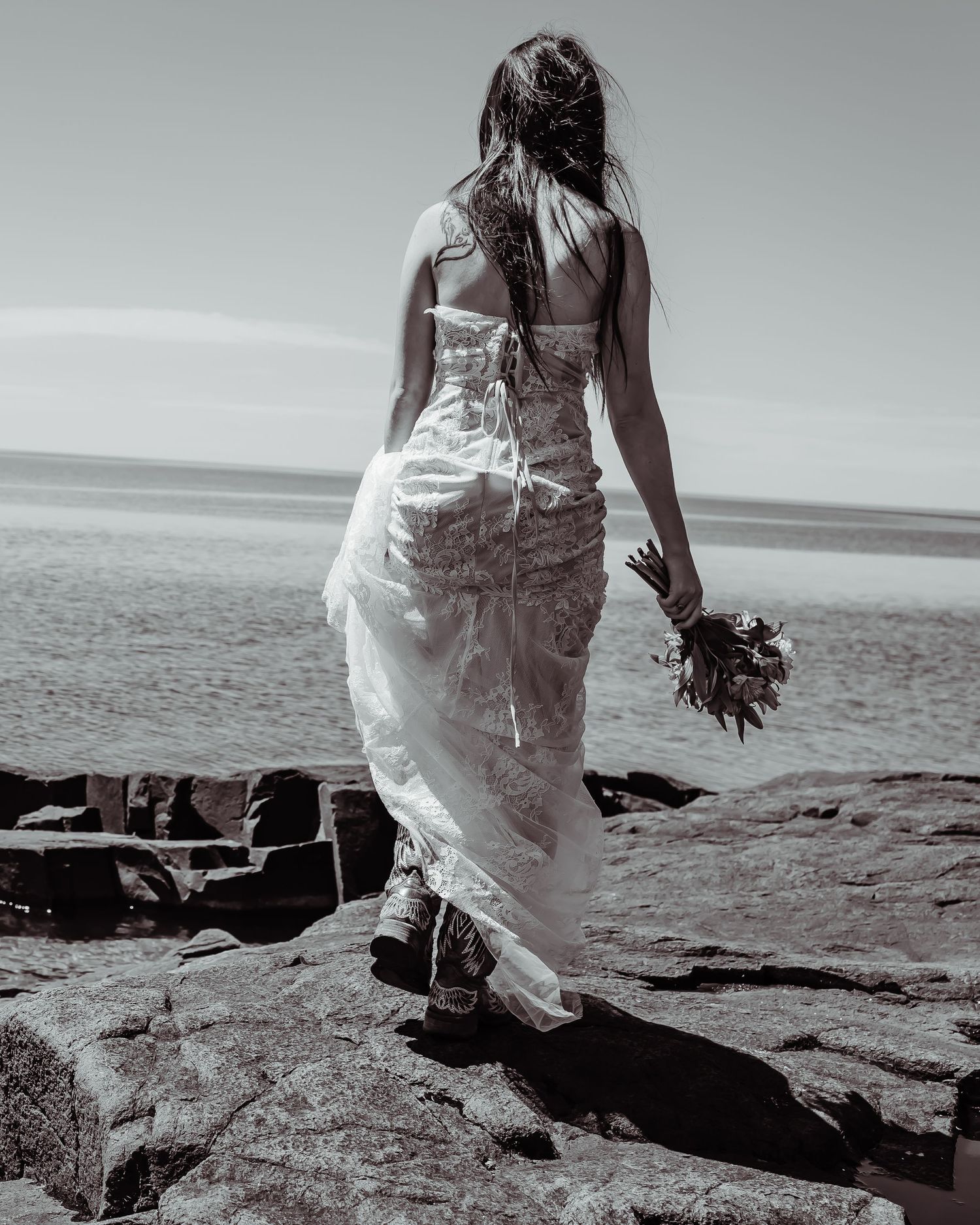 Monochrome image of bride in flowing dress standing on rocky beach while holding bouquet in black and white.
