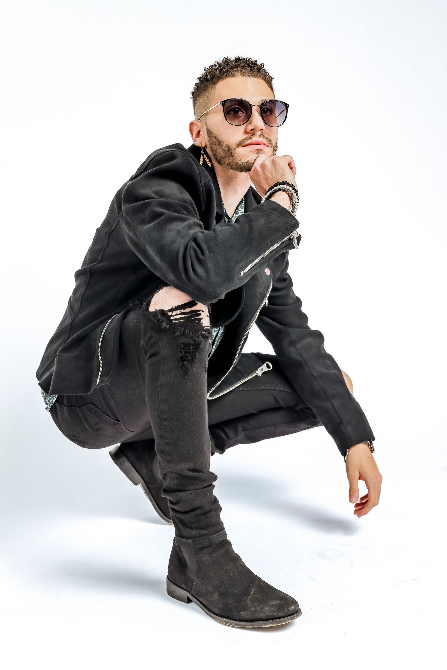 Stylish person in black leather jacket and sunglasses crouching in artistic pose against white background.