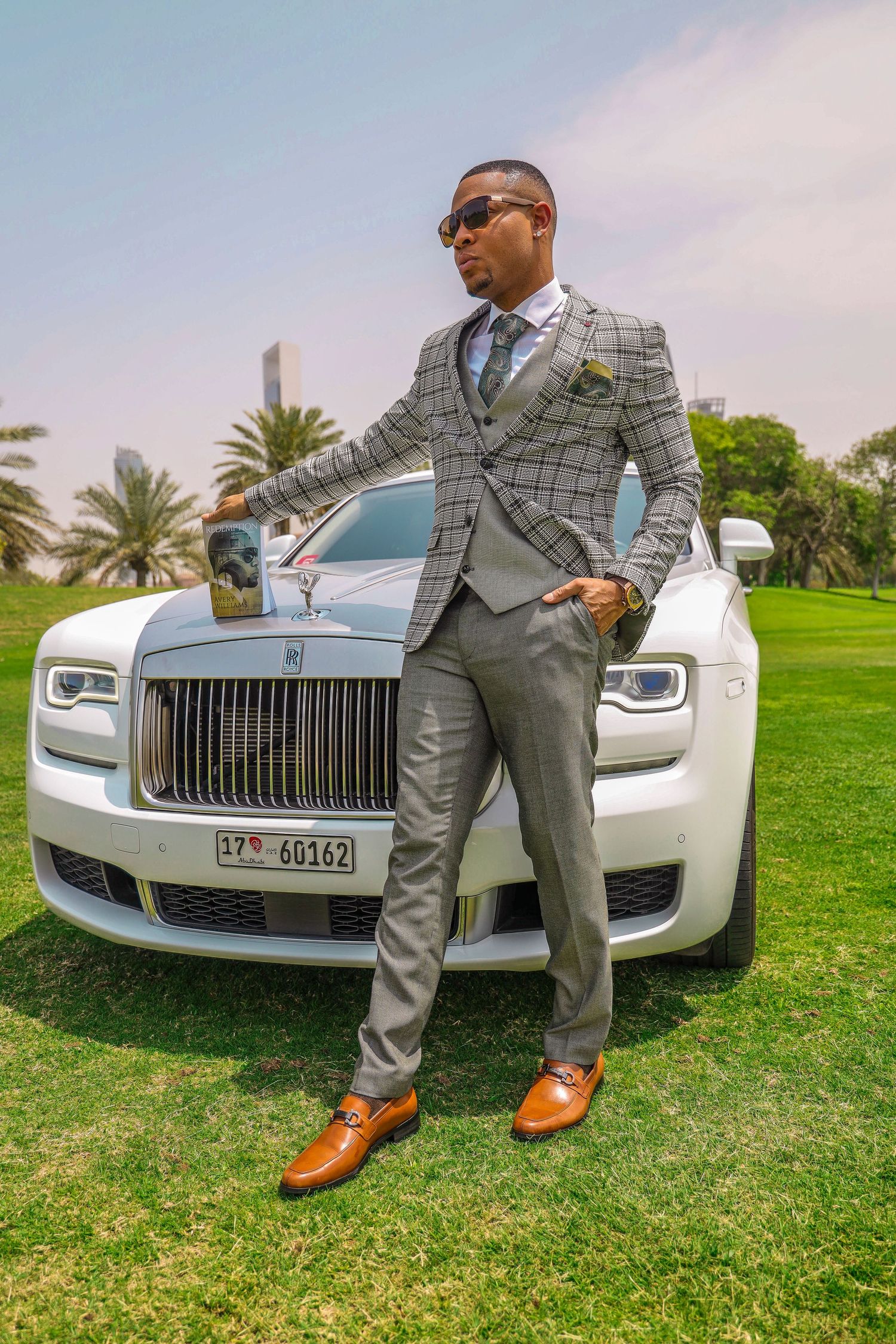 Luxury car enthusiast poses with white Rolls-Royce on manicured lawn with palm trees.