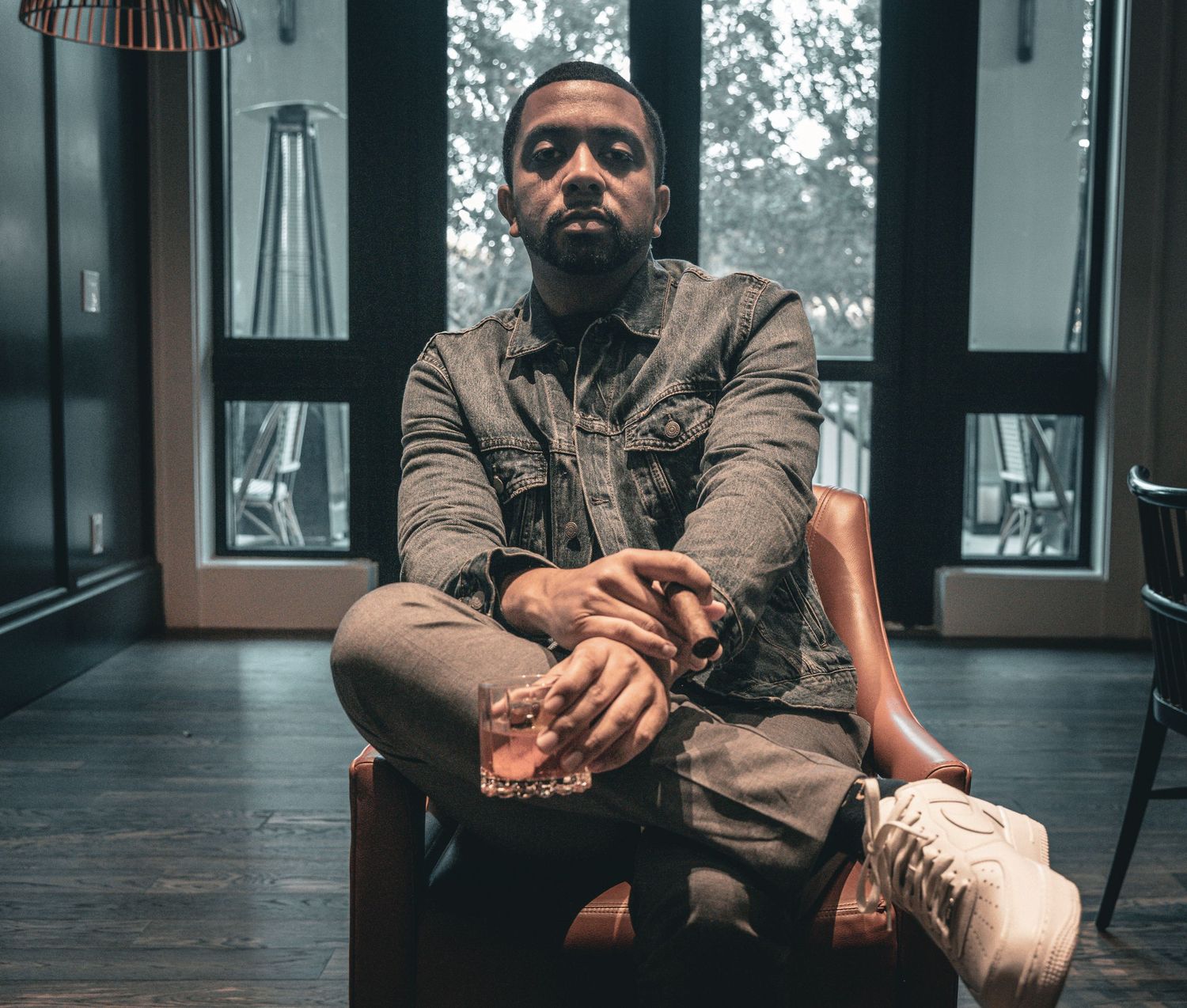 Person in casual attire sitting cross-legged on chair near windows with moody lighting.
