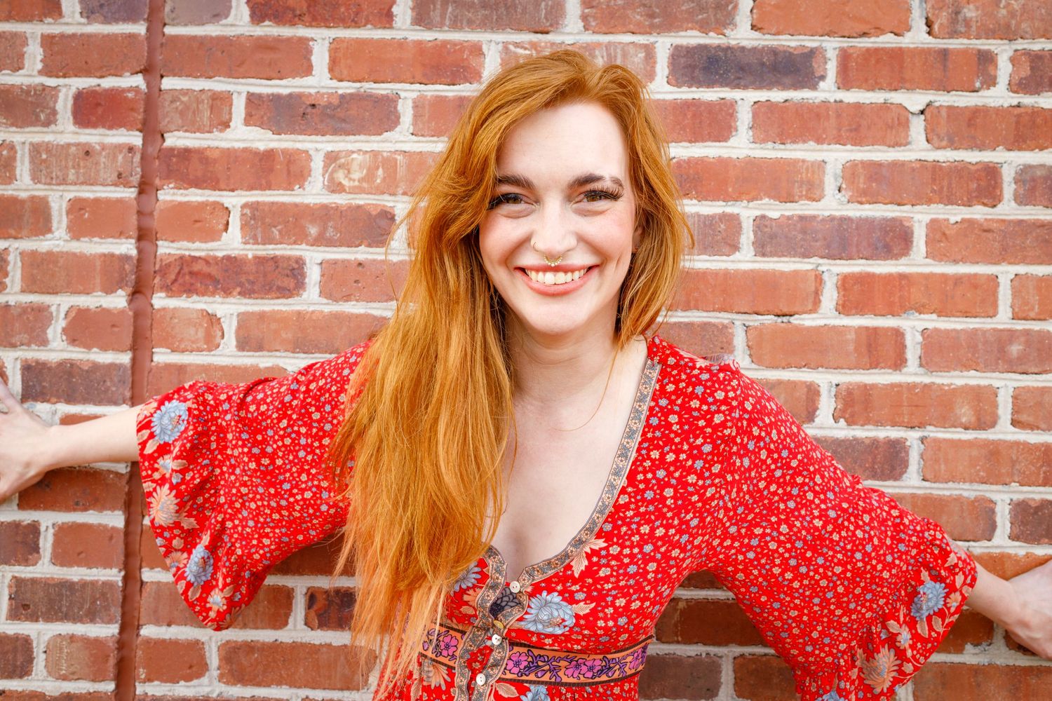 Woman with long red hair in red floral dress smiles against brick wall background.